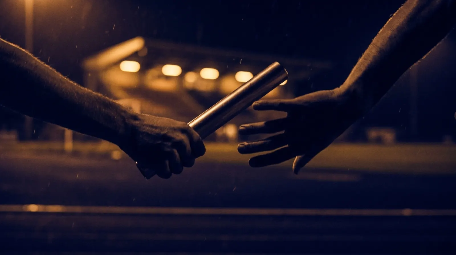 A relay baton handoff frozen mid-air under warm stadium lights.