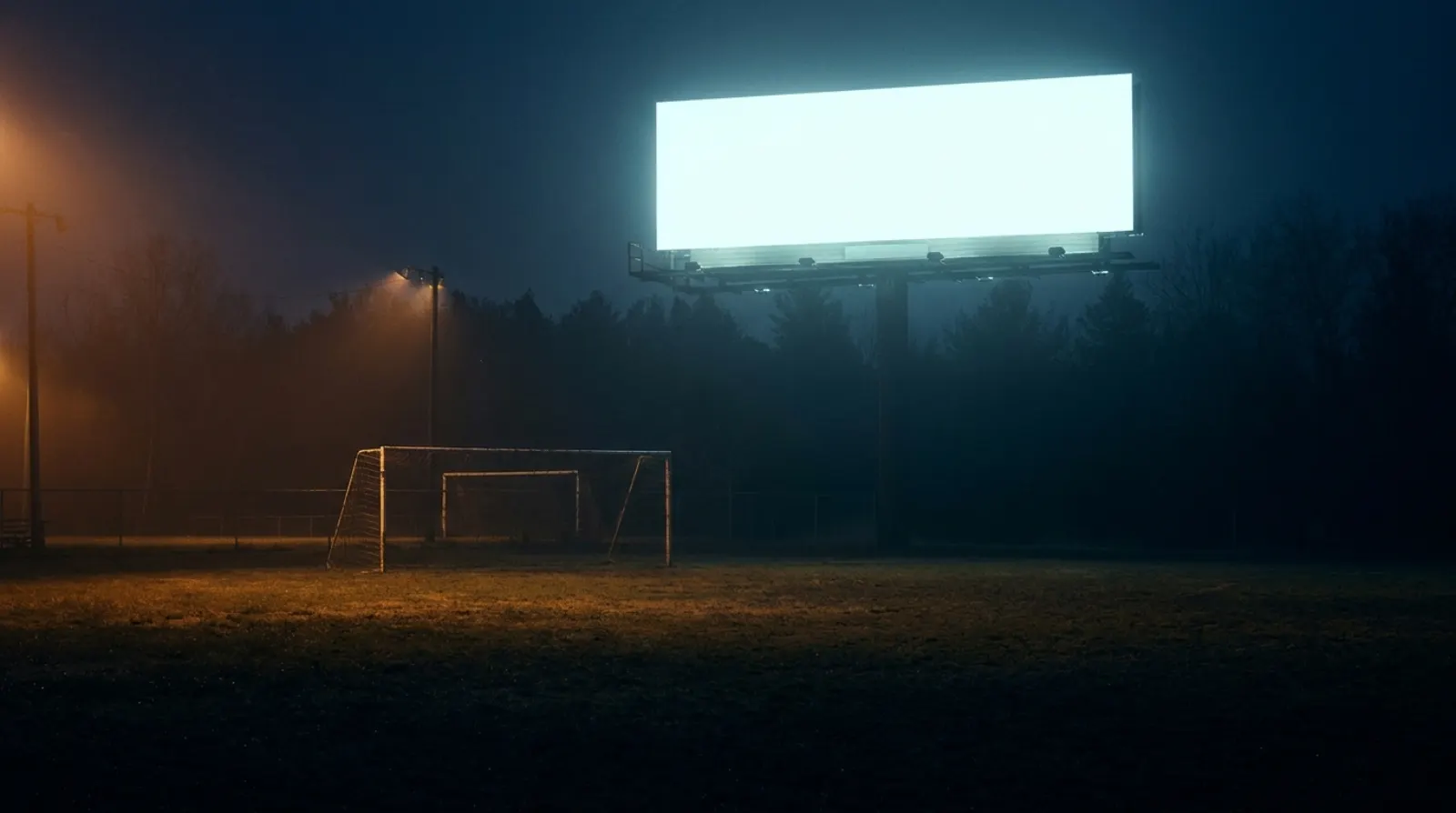 A huge blank billboard looming over an empty youth sports field at dusk.